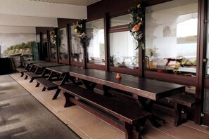 Une rangée de tables de pique-nique en bois, avec des bancs, de grandes fenêtres ornées de motifs floraux et une vue sur l’extérieur.