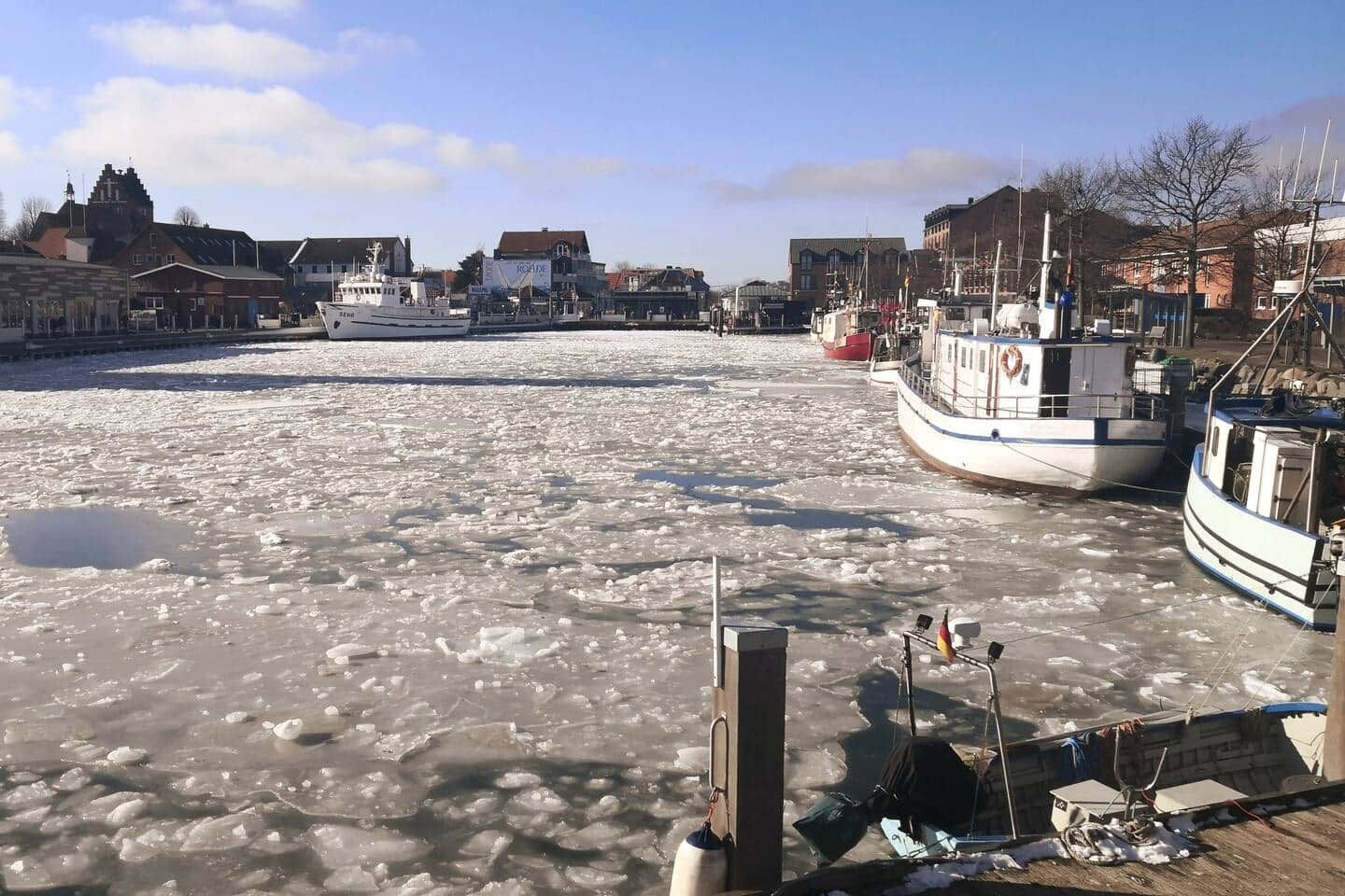 A frozen river with boats docked on the right, buildings in the background, and a clear sky.