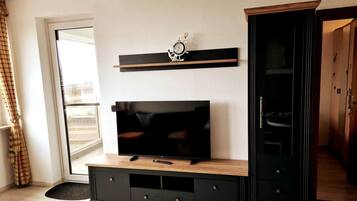 Ein Wohnzimmer mit Fernseher auf Holzständer, Regal darüber und einem Kleiderschrank zur Rechten.