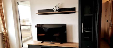 A living room with a TV on a wooden stand, a shelf above the TV, and a wardrobe to the right.