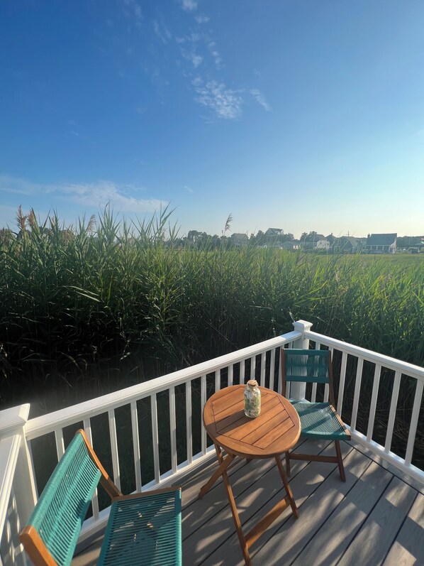 A wooden deck with a table and chairs, overlooking a field of tall grass and houses in the distance.