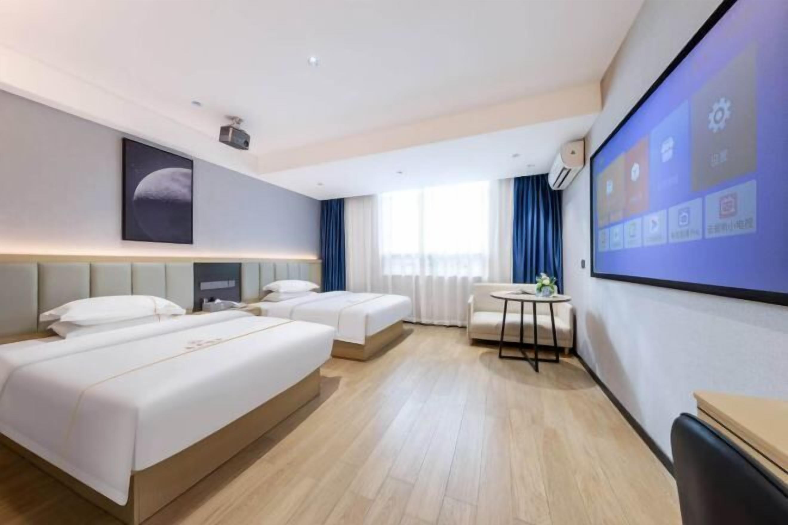 Habitación de hotel con tres camas, un escritorio, una silla, una pantalla para proyector y un ventanal con cortinas.