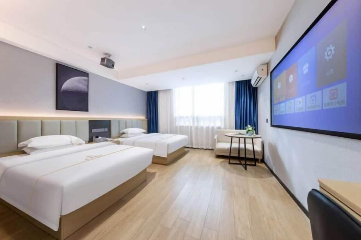 A hotel room with three beds, a desk, a chair, a projector screen, and a large window with curtains.