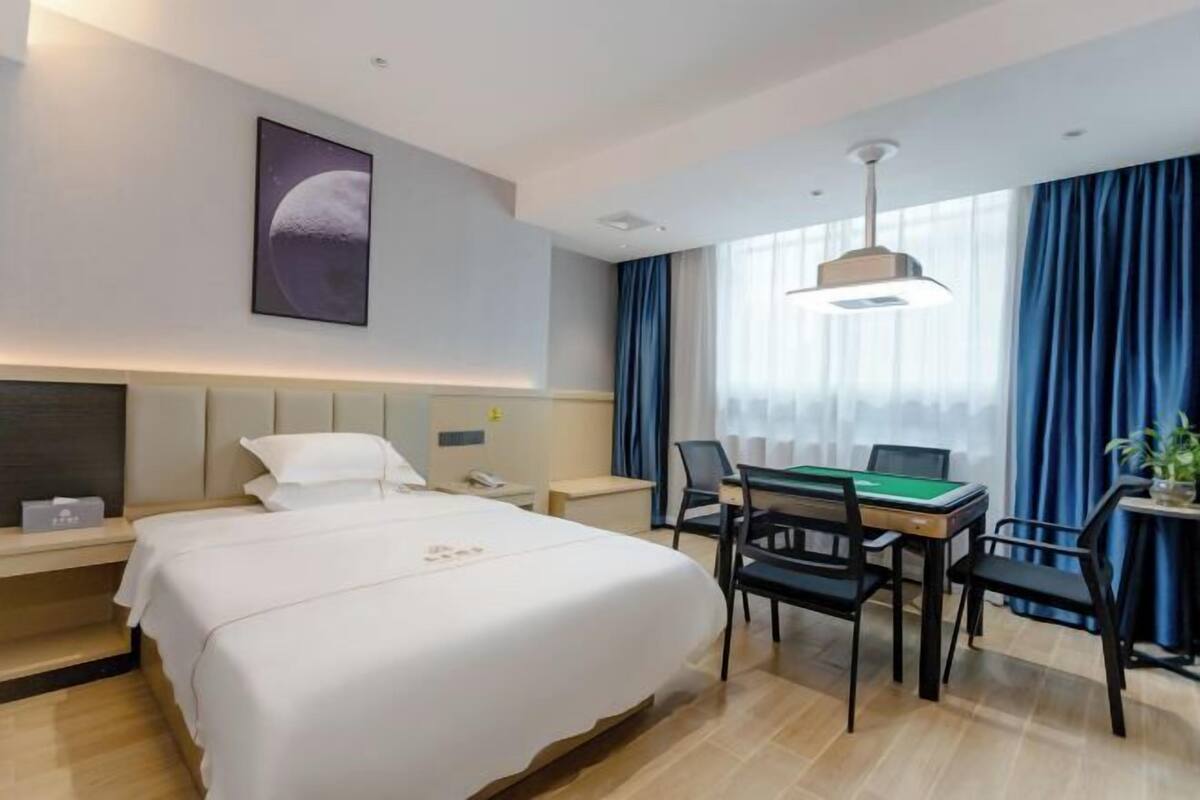 A hotel room with a large bed, a desk with a green felt table, and a wall-mounted picture of a planet.