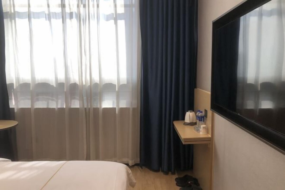 A hotel room with a bed, a small table with a kettle and cups, a TV, and a window with curtains.
