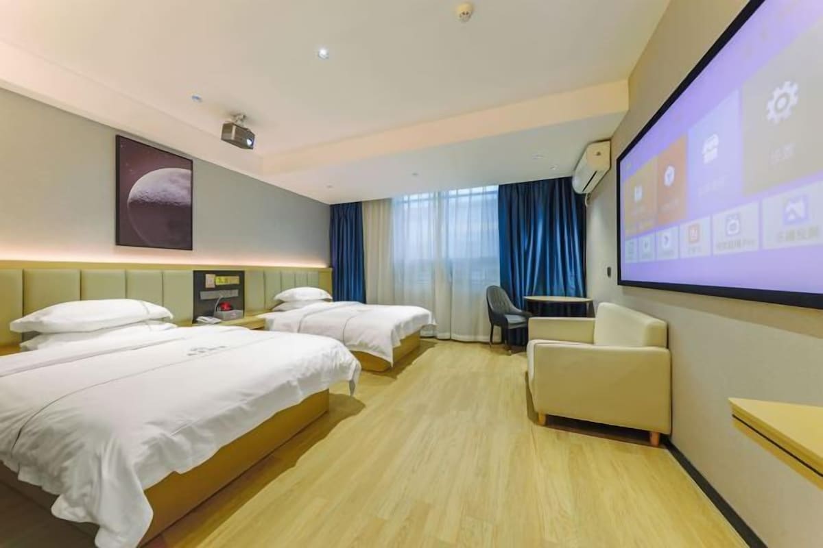 A hotel room with two beds, a desk, a chair, a projector screen, and a large window with curtains.