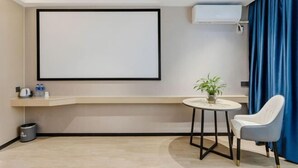 A modern hotel room with a large flat-screen TV mounted on the wall, a small round table with a potted plant, a white chair, and a blue curtain.