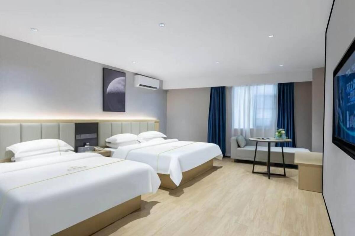 A hotel room with two beds, a small table, a sofa, and a large window with curtains.