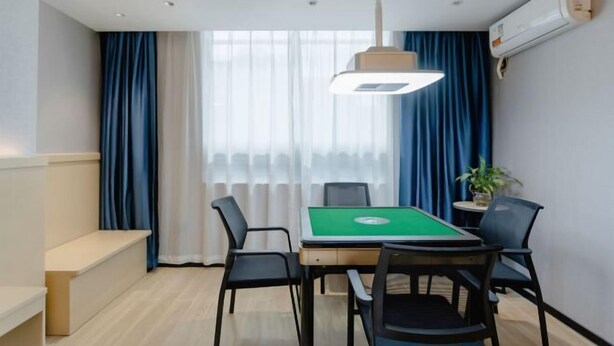 A room with a green pool table, black chairs, a bench, a hanging light fixture, and a window with blue curtains.