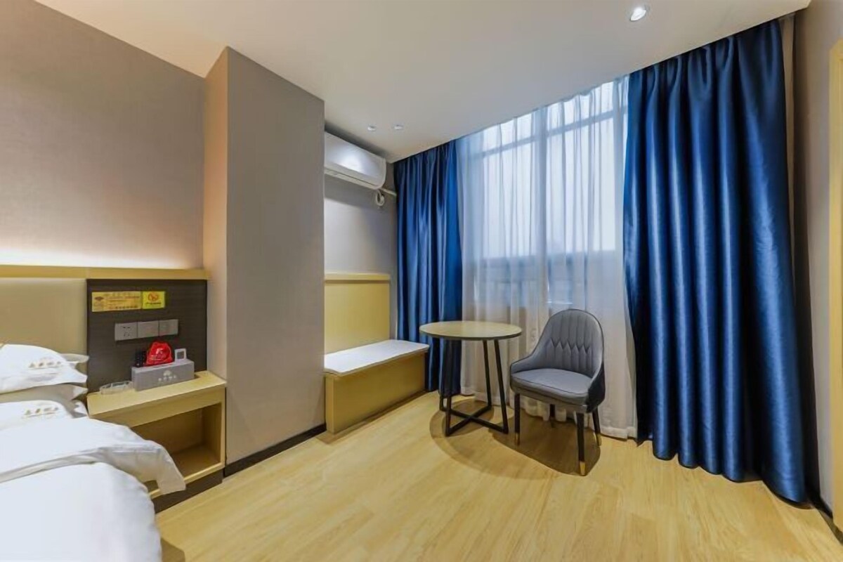 A hotel room with a bed, a small table, a chair, a bench, and a window with blue curtains.
