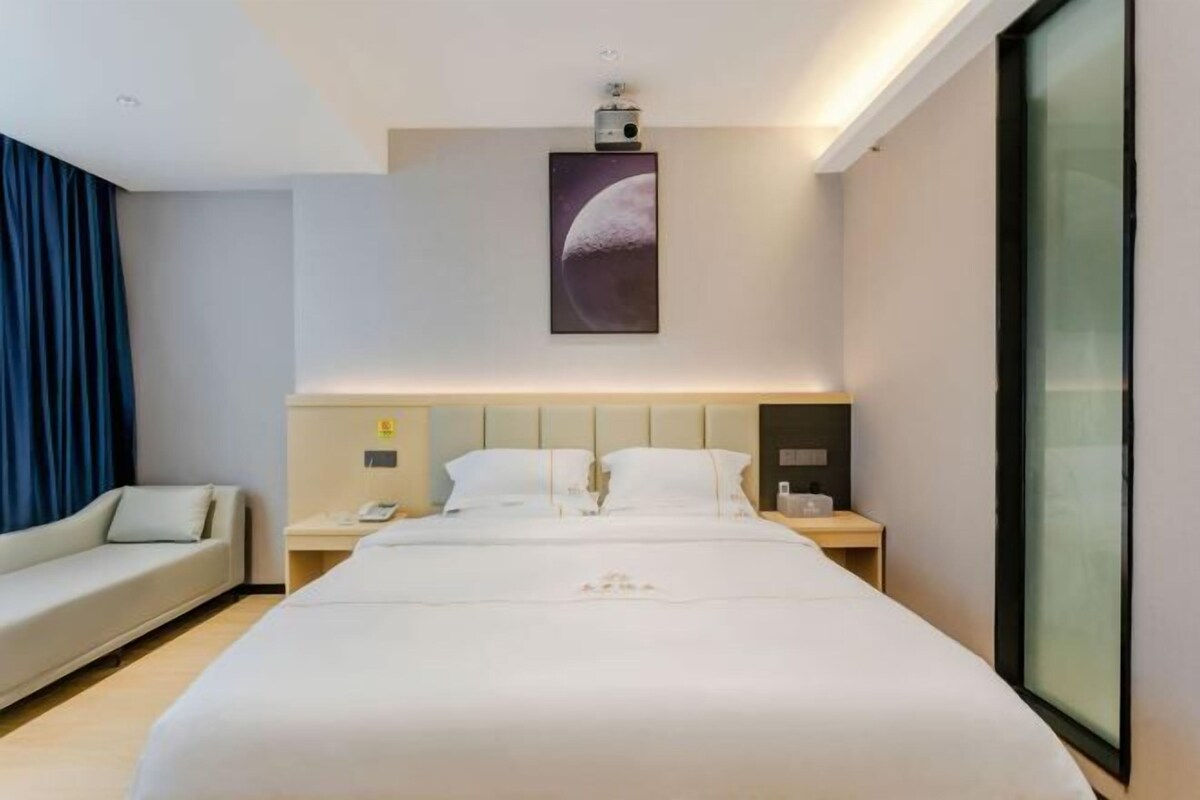 A modern hotel room with a large bed, a bedside table with a lamp, a nightstand, a sofa, and a large window.