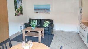 Ein Wohnzimmer mit einer Couch, einem Couchtisch und einem Esstisch. Auf dem Tisch stehen Weingläser und eine Weinflasche. An den Wänden hängen zwei Bilder, eines zeigt eine Straße, das andere einen Strand.