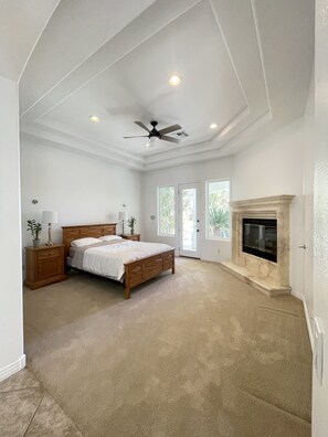 A bedroom with a large bed, a ceiling fan, a fireplace, and a door leading to a balcony.