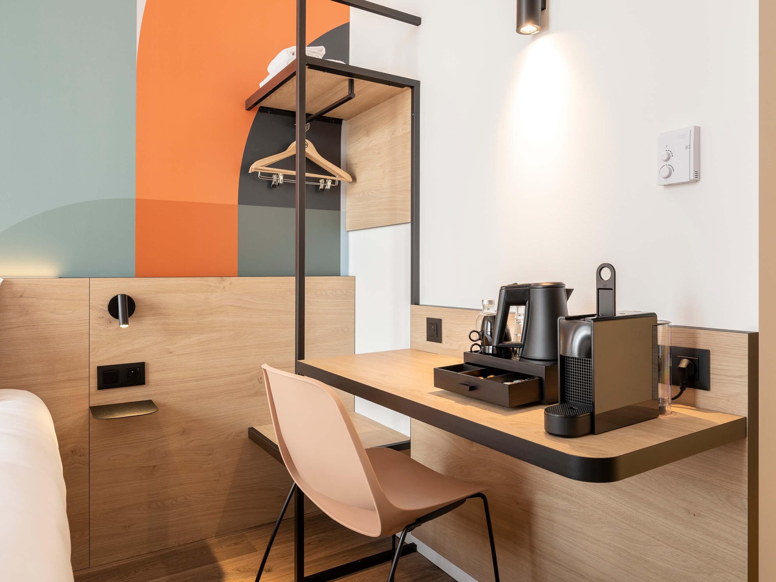 Una habitación de hotel moderna con un escritorio de madera, una silla, una cafetera y un perchero.