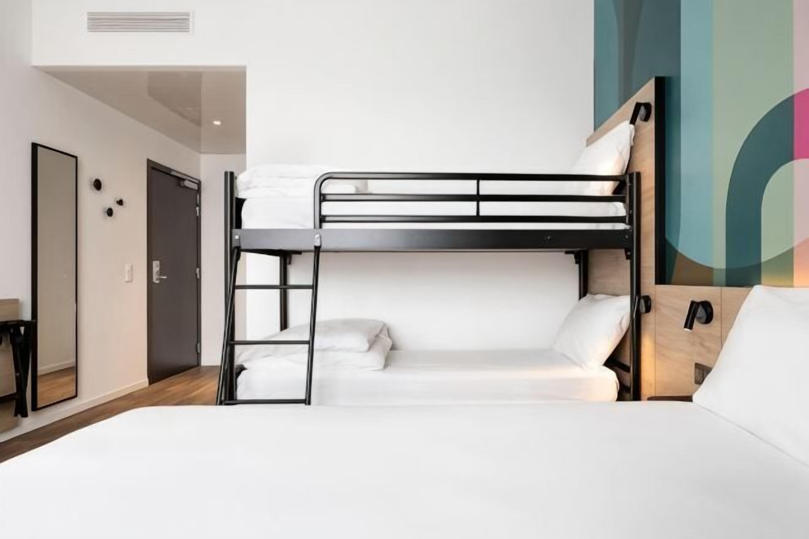 A bunk bed in a hotel room with a mirror, a door, and a wall with abstract art.