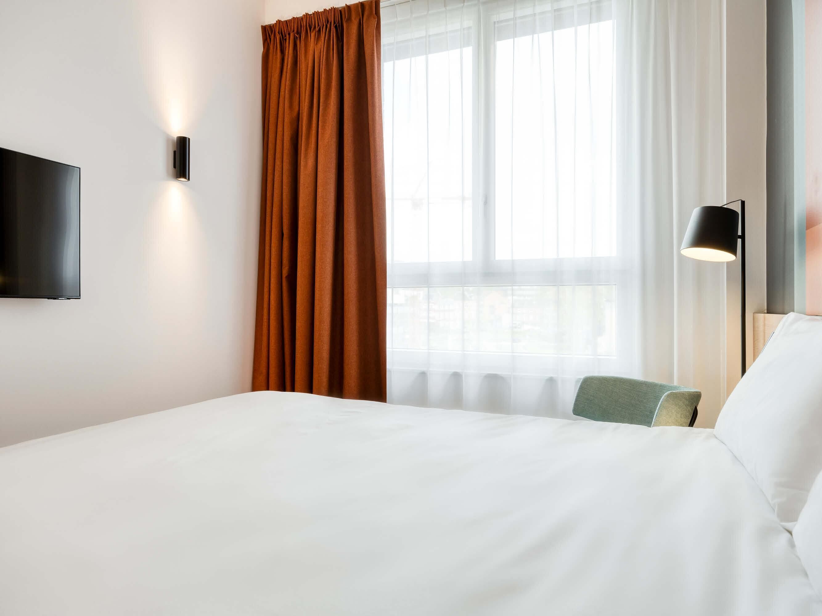 A hotel room with a bed, a TV, a window with sheer curtains, and a wall-mounted lamp.