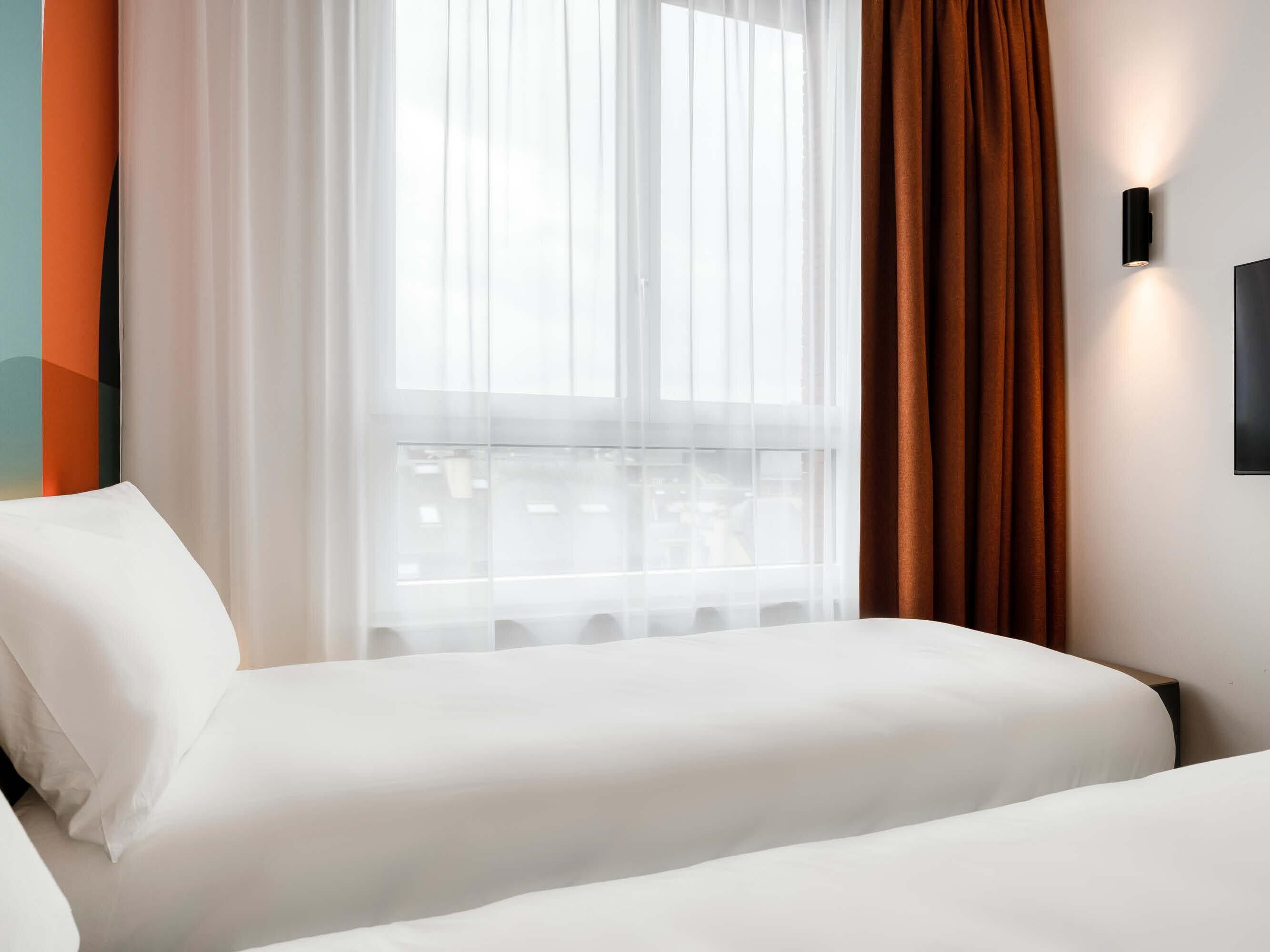 Une chambre d’hôtel avec un lit, une fenêtre avec des rideaux légers et un luminaire fixé au mur.