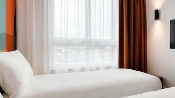Ett hotellrum med en säng, ett fönster med tunna gardiner och en väggmonterad lampa.