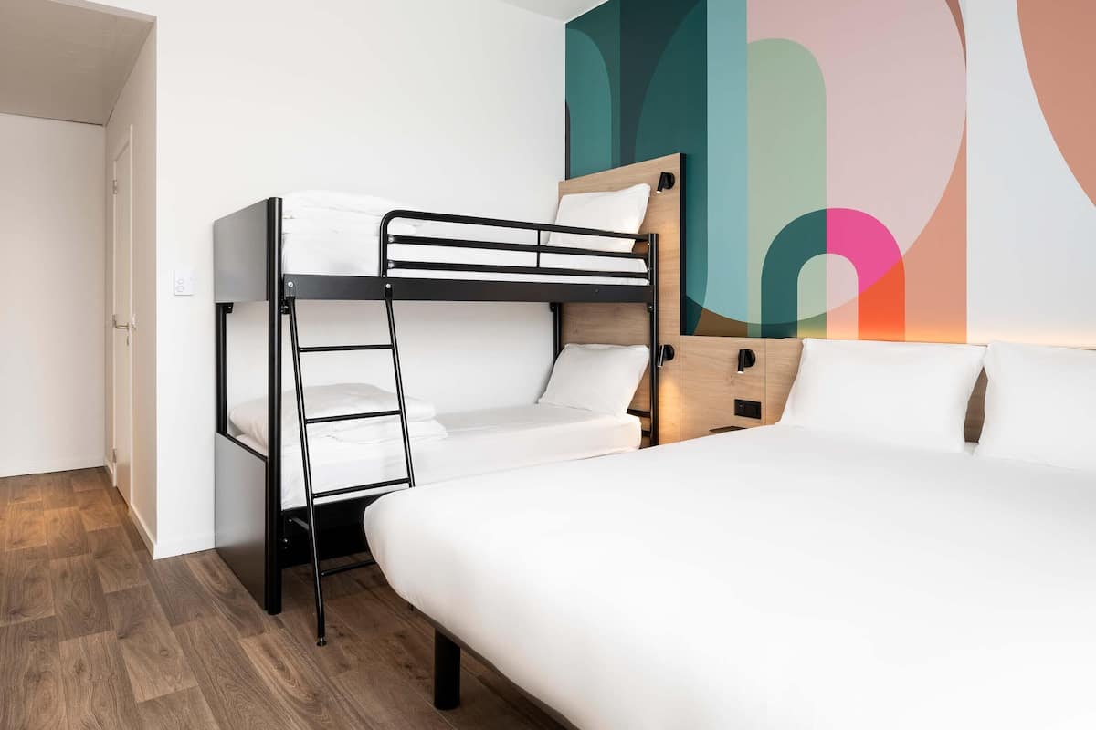 Een slaapkamer met een stapelbed, een houten hoofdbord, een wit matras en kleurrijke abstracte muurkunst.