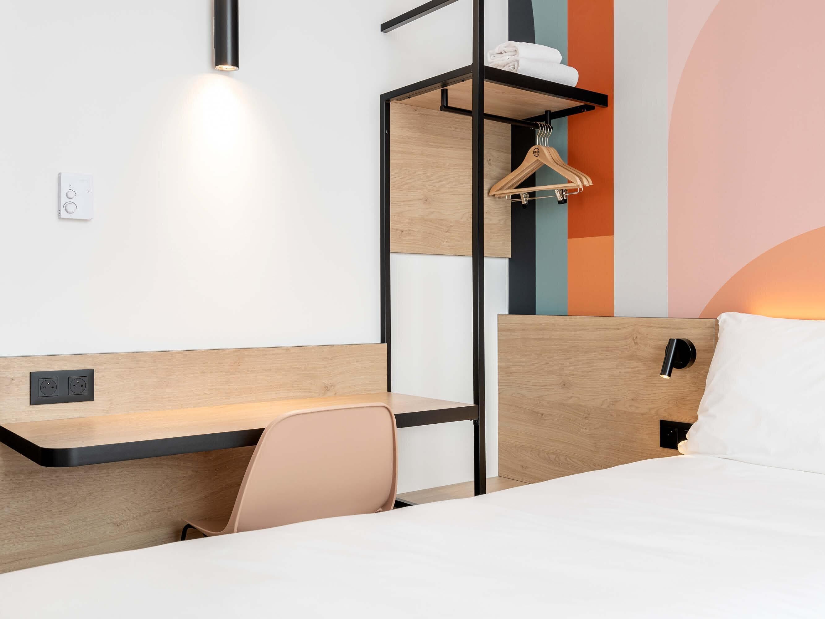 A hotel room with a bed, a desk, a chair, a hanging coat rack, and a wall-mounted light fixture.