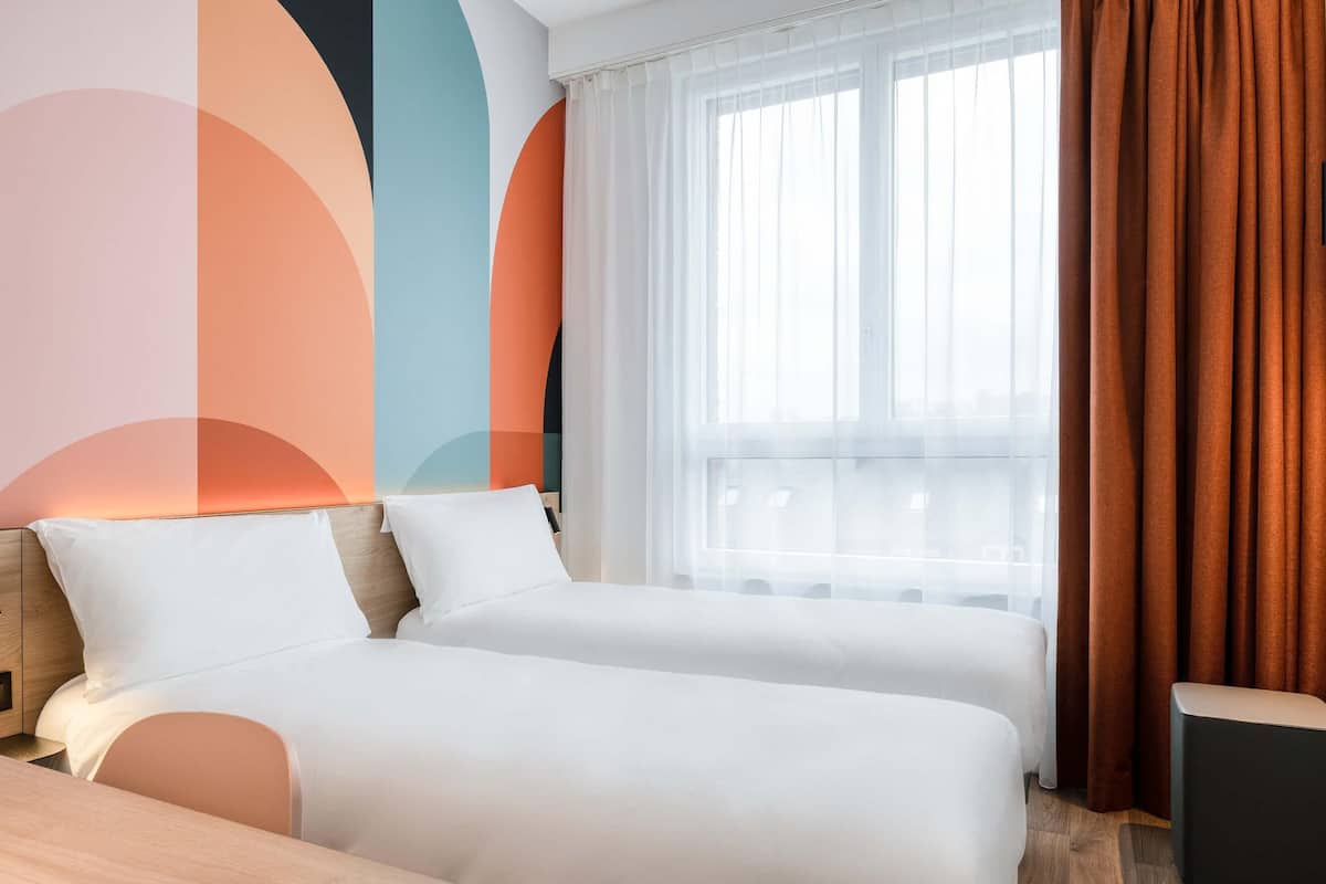 Een hotelkamer met twee bedden, een houten hoofdbord, een raam met dunne gordijnen en uitzicht op de stad.