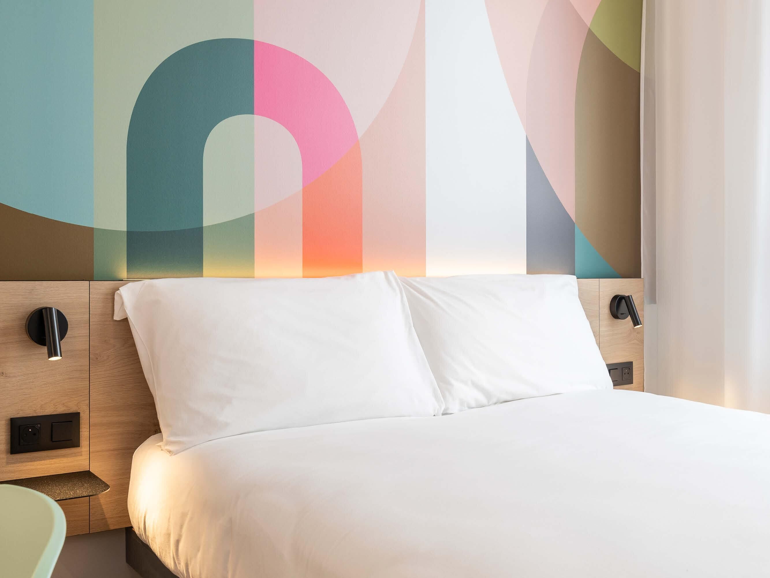 A modern hotel room with a wooden headboard, a bed with white linens, a nightstand, and a wall-mounted lamp.
