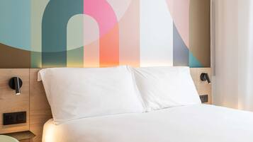 A modern hotel room with a wooden headboard, a bed with white linens, a nightstand, and a wall-mounted lamp.