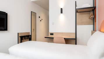 A modern hotel room with a bed, a desk, a chair, a TV, and a shelf with towels.