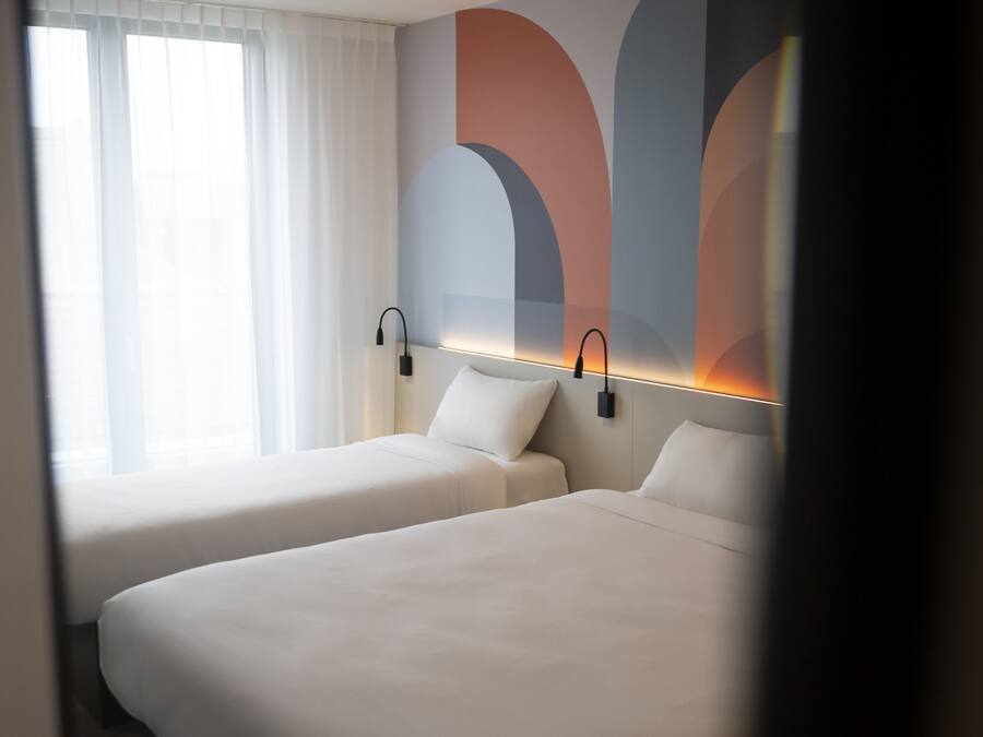 Een hotelkamer met twee bedden, een groot raam met witte gordijnen, en een muur met abstracte kunst.