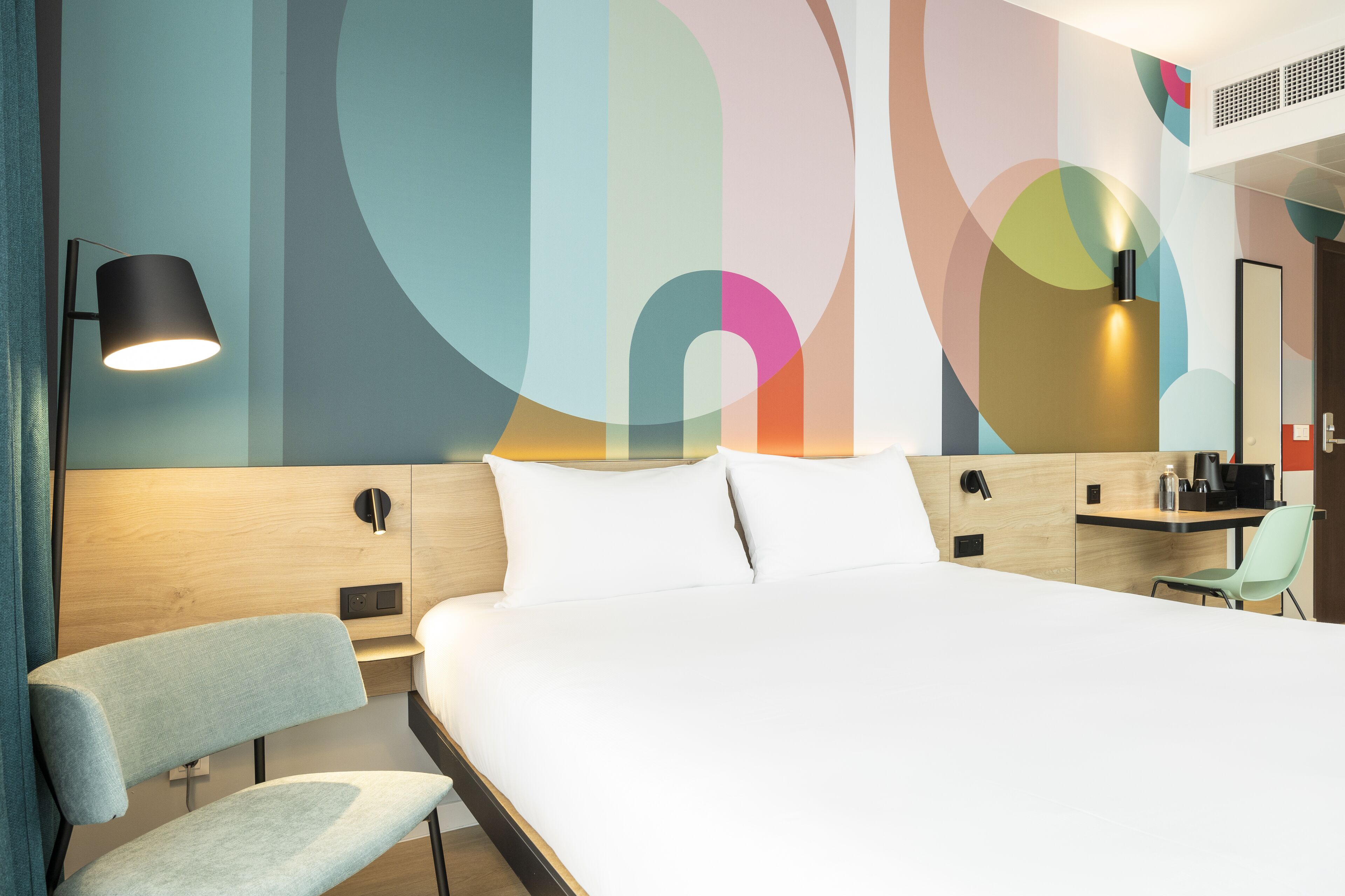 Et moderne hotelværelse med en stor seng, et skrivebord, en stol og en væg med abstrakt kunst.