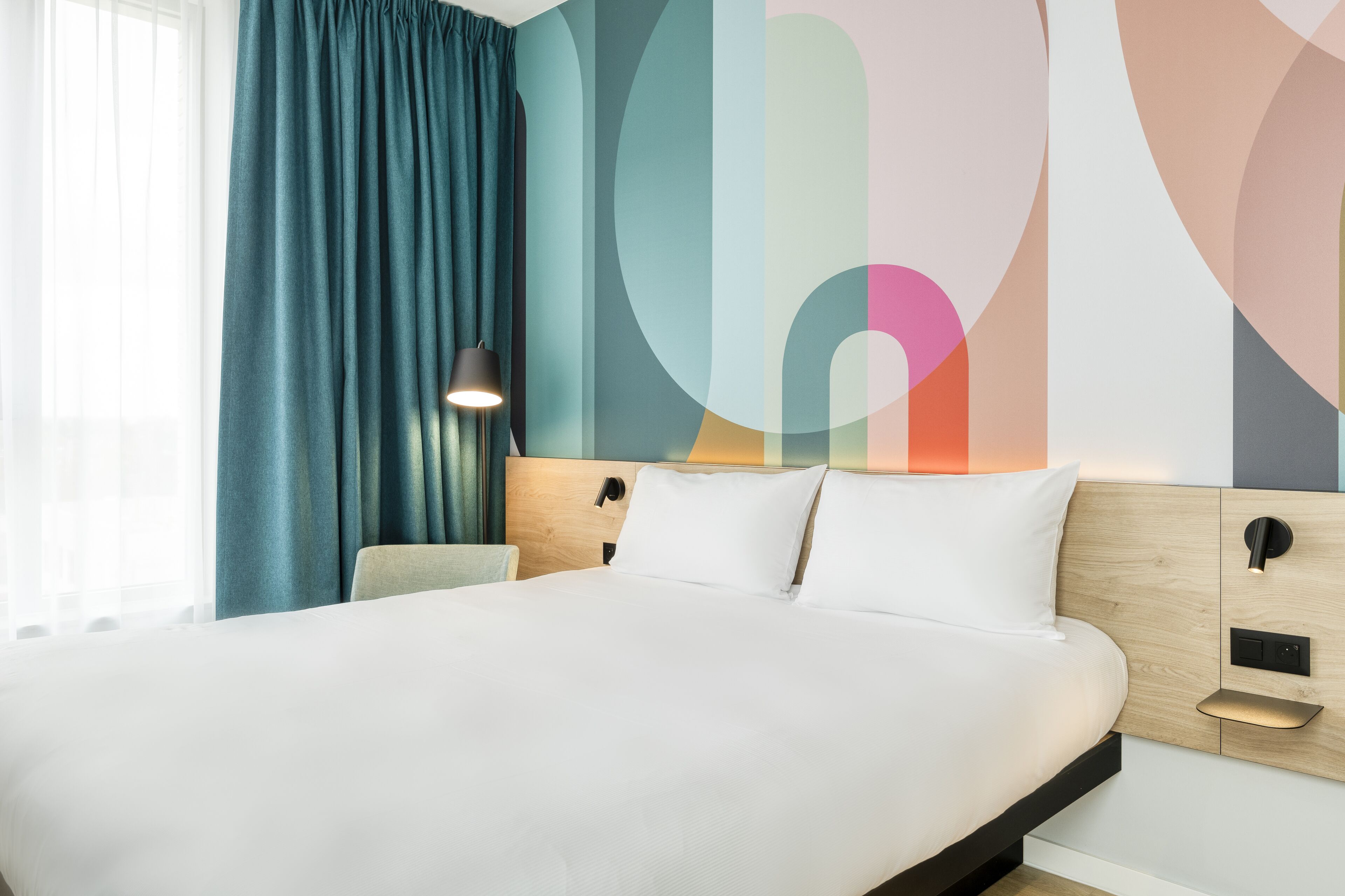 Ein modernes Hotelzimmer mit einem großen Bett, einem Nachttisch, einer Lampe und einer Wand mit abstrakter Kunst.