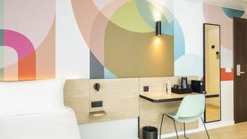 A modern hotel room with a bed, a desk with a chair, a mirror, and a trash bin.