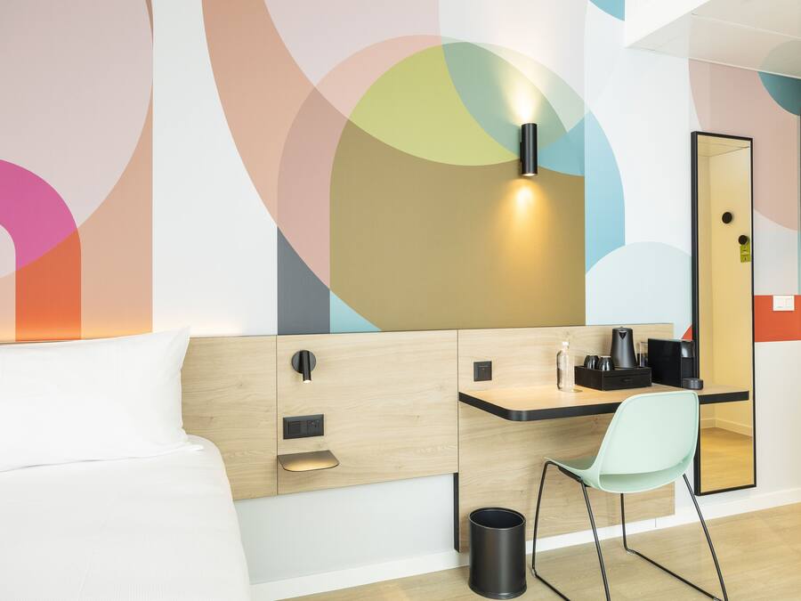A modern hotel room with a bed, a desk with a chair, a mirror, and a trash bin.