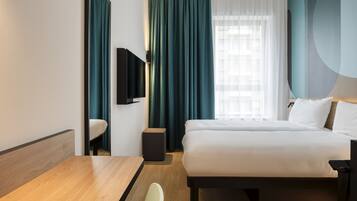 A hotel room with a bed, a desk, a chair, a TV, and a window with curtains.
