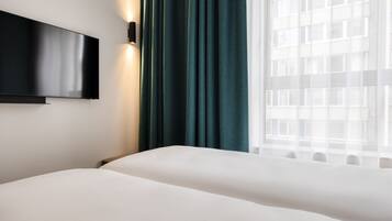 Une chambre d’hôtel dotée d’un téléviseur à écran plat fixé au mur, d’un lit avec une literie blanche, de rideaux turquoise et d’une fenêtre donnant sur un immeuble.