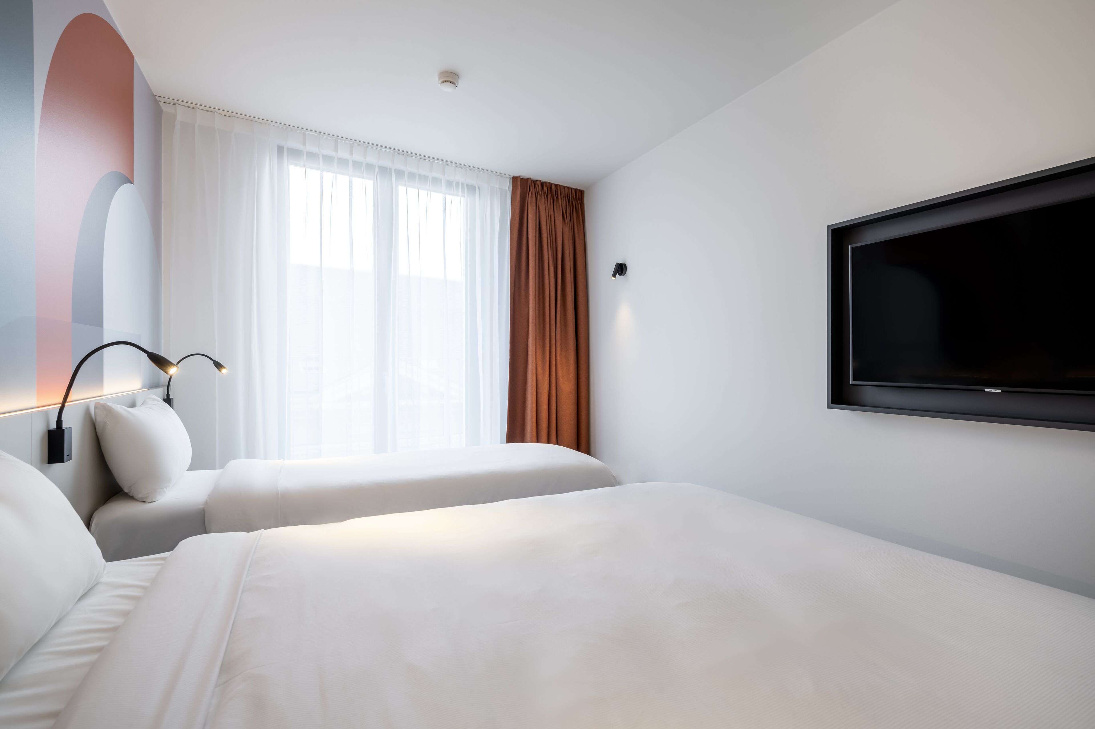 Habitación de hotel con dos camas, un ventanal grande con cortinas translúcidas, un televisor montado y una lámpara de pared.