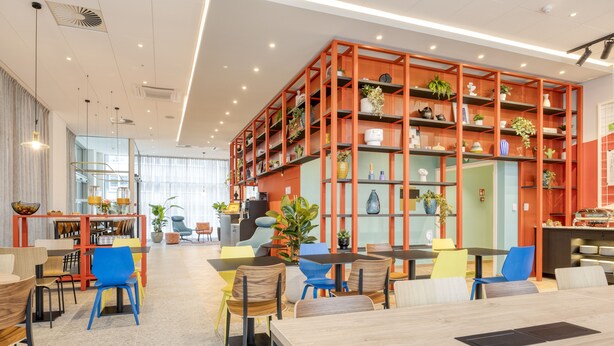 Un comedor moderno con mesas de madera y sillas coloridas, rodeado por una estantería roja y vibrante llena de plantas y objetos decorativos.