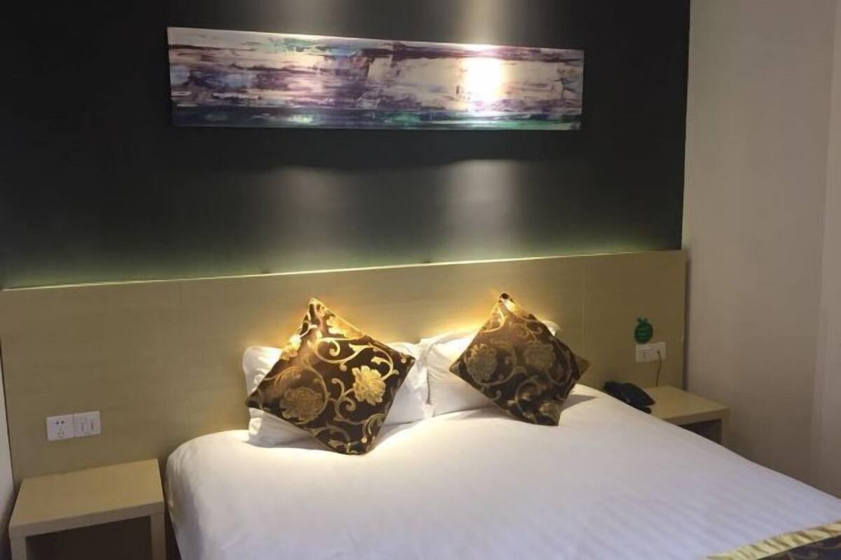 Habitación de hotel con cama, dos almohadones decorativos, mesita de noche, teléfono y un cuadro colgado en la pared.