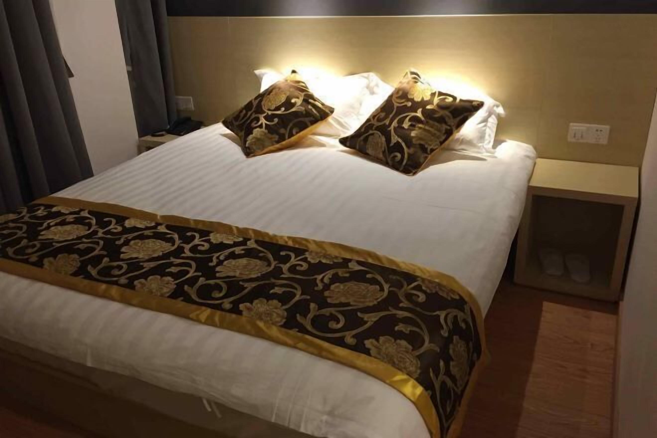 Un lit bien fait, recouvert d’une couvre-lit à motifs et agrémenté de deux coussins décoratifs. On y trouve une table de chevet avec une lampe et une petite étagère où sont posées des chaussons.