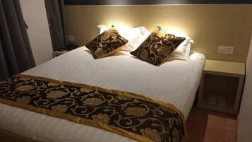 Un lit bien fait, recouvert d’une couvre-lit à motifs et agrémenté de deux coussins décoratifs. On y trouve une table de chevet avec une lampe et une petite étagère où sont posées des chaussons.