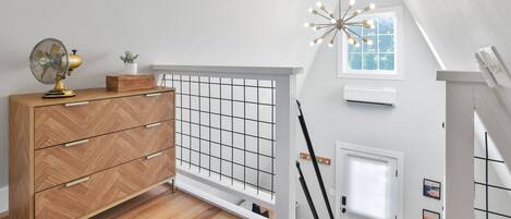 A modern loft bedroom with a wooden dresser, a fan, a grid-patterned window, and a unique ceiling light fixture.