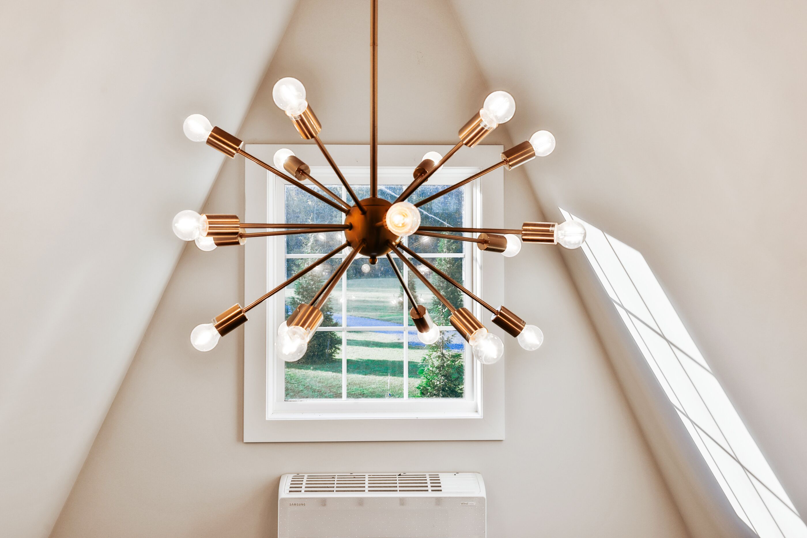 Ein moderner Kronleuchter mit mehreren Lampen, ein Fenster mit Blick auf Grünflächen und eine weiße Klimaanlage.