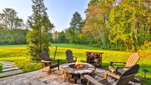 A circular stone fire pit surrounded by Adirondack chairs, set on a stone patio with a grassy field and trees in the background. - Escape to this unique a-frame cabin minutes from Old Rag Mountain  (Etlan)