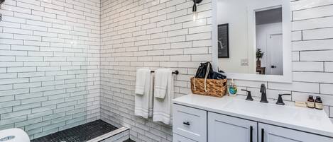 Un baño moderno con azulejos blancos tipo metro, un área de ducha hexagonal negra, un inodoro blanco y un tocador blanco con espejo.