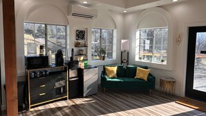 A modern kitchen with a built-in microwave, coffee machine, and refrigerator. There are two windows providing natural light and a view of the outside. A green sofa with gold pillows is placed in the living area. - The Four Daughters (Oakhurst)