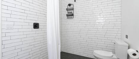 A bathroom with white brick walls, a white toilet, a black trash bin, and a shower curtain.