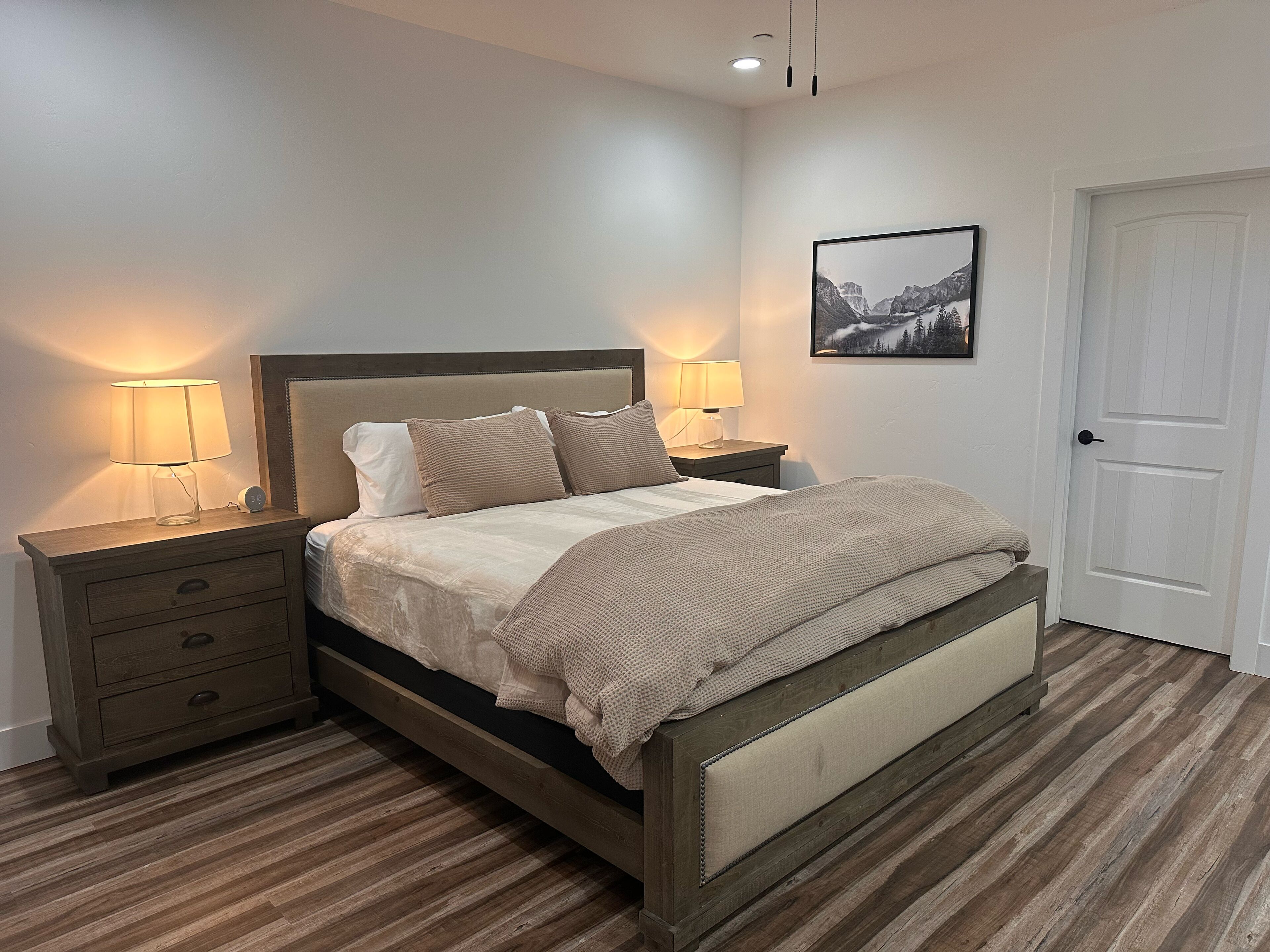 Un dormitorio ordenado con cama, dos mesitas de noche con lámparas, una puerta y un cuadro enmarcado en la pared.