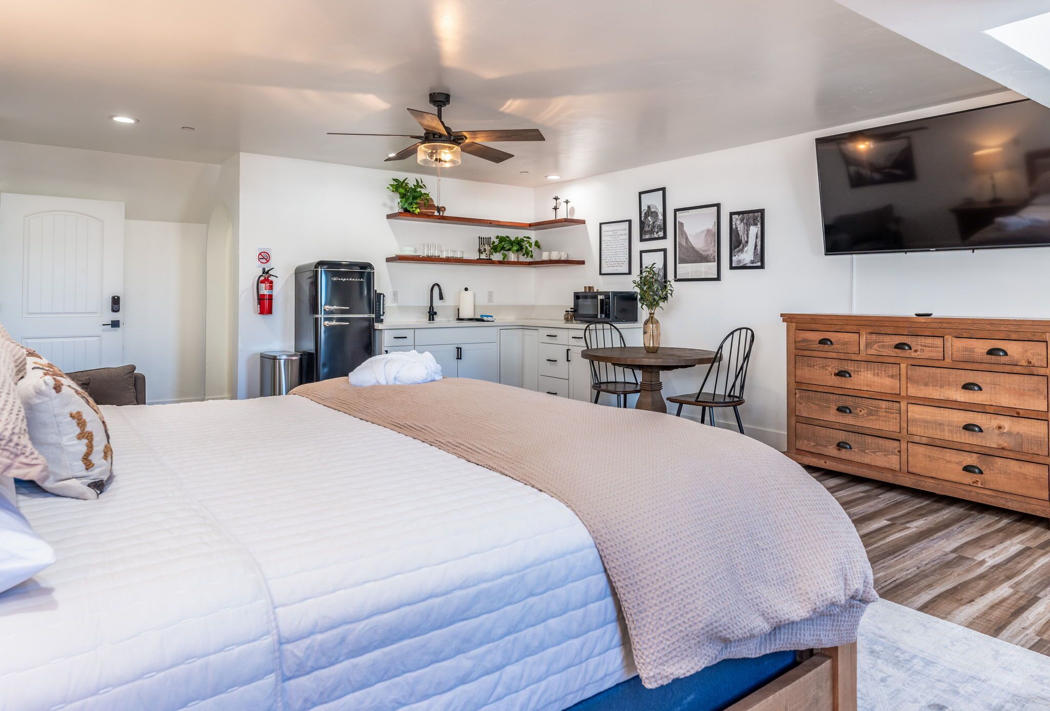 A modern bedroom with a large bed, a ceiling fan, a TV, a kitchen area with a fridge, microwave, and a dining table.