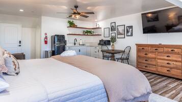 A modern bedroom with a large bed, a ceiling fan, a TV, a kitchen area with a fridge, microwave, and a dining table.