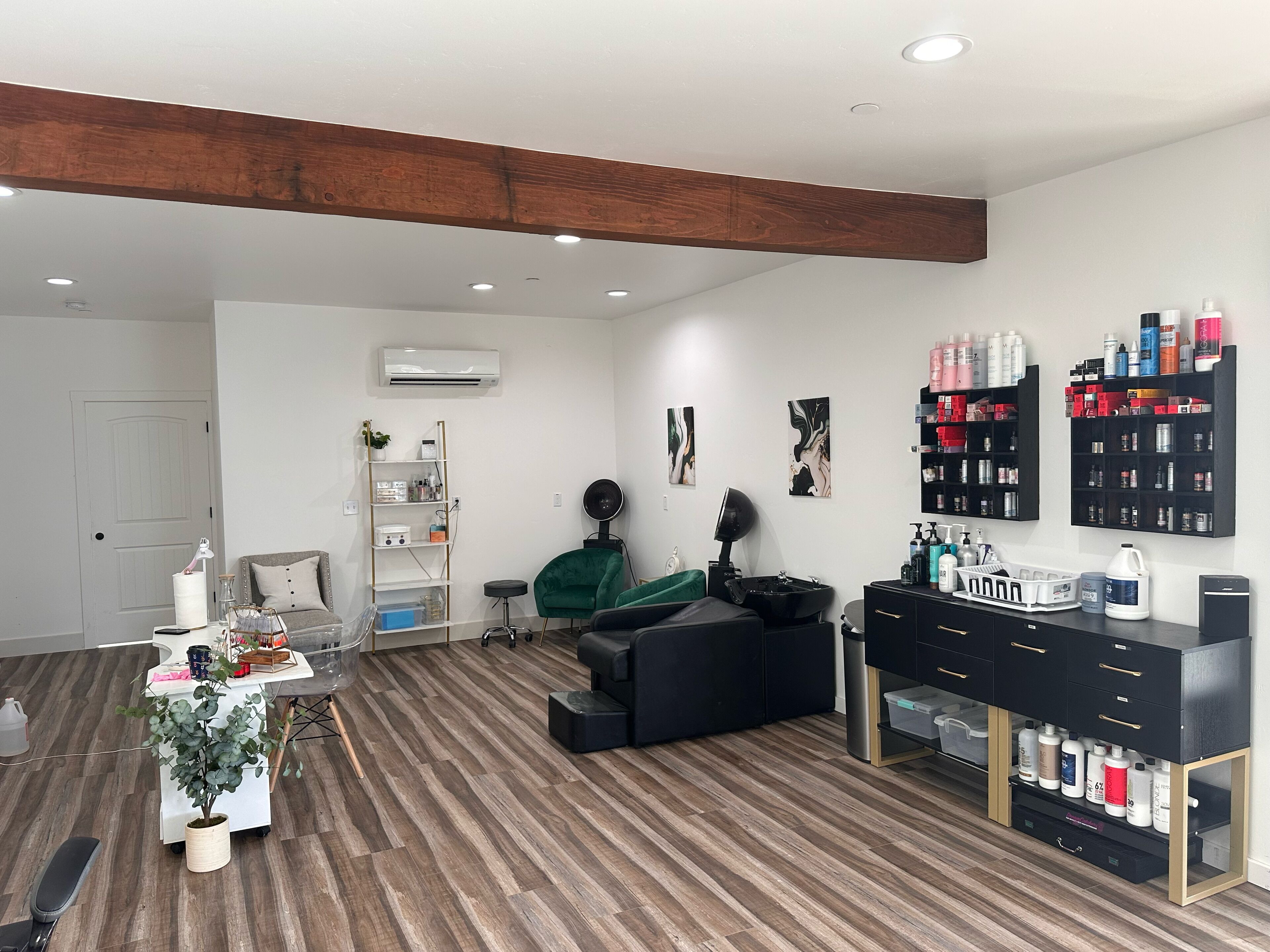 Un salón con suelo de madera, un sofá negro, una silla verde y estanterías con productos para el cabello.
